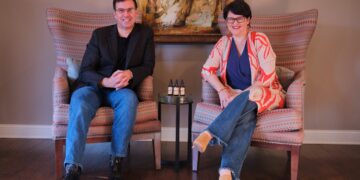 A man in a black blazer and a woman in a patterned orange and white cardigan sit in large patterned wingback chairs. They smile at the camera, with a small round glass table holding three dropper bottles positioned between them.