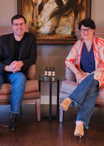 A man in a black blazer and a woman in a patterned orange and white cardigan sit in large patterned wingback chairs. They smile at the camera, with a small round glass table holding three dropper bottles positioned between them.