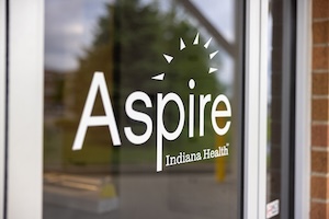 A close-up of the Aspire Indiana Health logo in white vinyl on a glass door. The logo features the word "Aspire" with a stylized sunburst above the letter "i" and "Indiana Health" written in smaller text below. The background through the glass is softly blurred, showing an outdoor area.