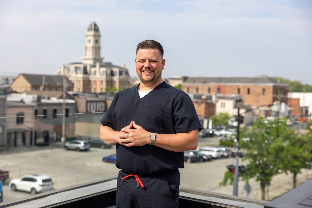 A medium shot of a man in black medical scrubs standing outdoors on a rooftop or balcony. He is smiling warmly with his hands clasped in front of him. Behind him, a blurred cityscape features historic buildings, a prominent clock tower, and a parking lot under a clear sky.