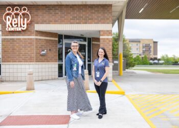 Two women standing and smiling outside the entrance of the Rely Center, which is part of Aspire Indiana Health. The building is made of red and tan brick. One woman wears a denim jacket and a long checkered skirt, while the other wears a blue Rely-branded polo shirt and black trousers. A large white "Rely Center" logo is visible on the brick wall behind them.