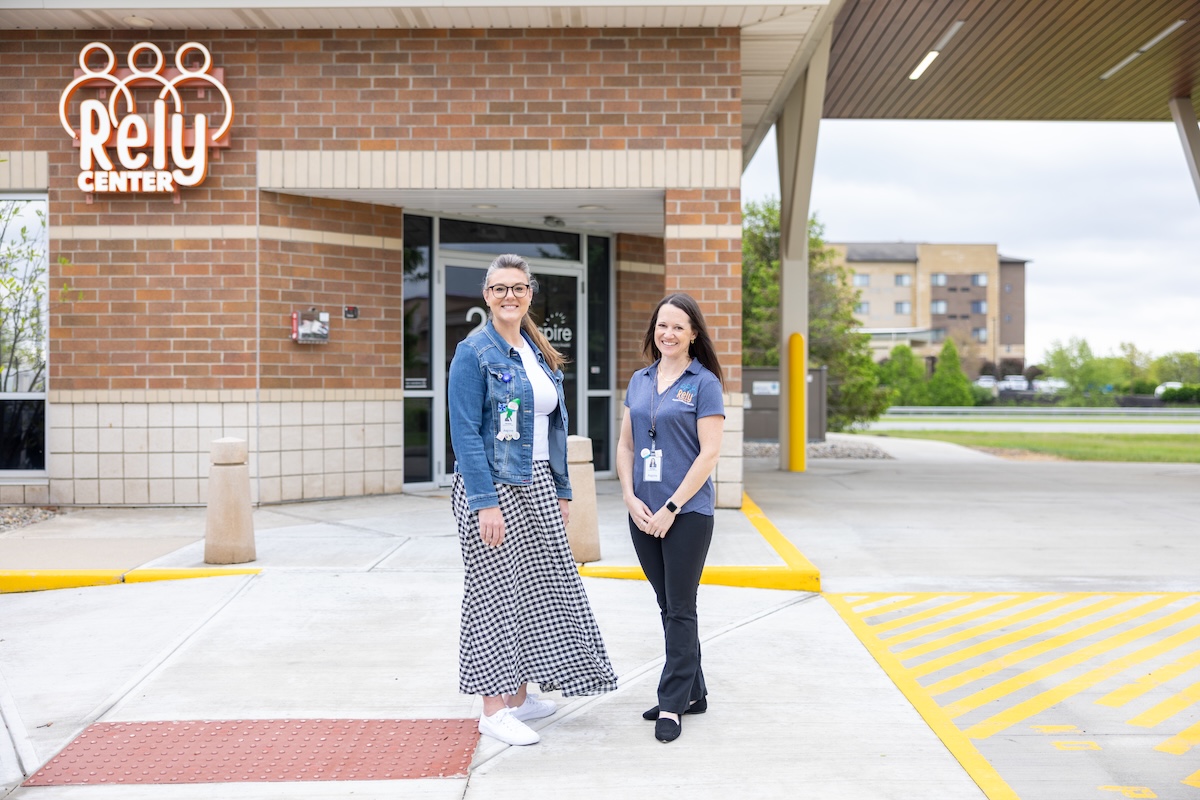 Two women standing and smiling outside the entrance of the Rely Center, which is part of Aspire Indiana Health. The building is made of red and tan brick. One woman wears a denim jacket and a long checkered skirt, while the other wears a blue Rely-branded polo shirt and black trousers. A large white "Rely Center" logo is visible on the brick wall behind them.