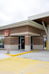 An exterior shot of the Rely Center entrance, featuring a modern brick building with a large "Rely Center" logo in white and orange. The entrance is marked with a large number "2" on a glass door. Bright yellow safety markings are painted on the concrete ground in front of the building under a wide overhang.