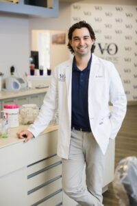 A professional portrait of an orthodontist, a man with dark hair and a smile, wearing a white lab coat over a navy polo and tan trousers. He is leaning against a light-colored counter in a clean, modern orthodontic office. The Volovic Orthodontics logo is visible on his lab coat and on the wall in the blurred background.