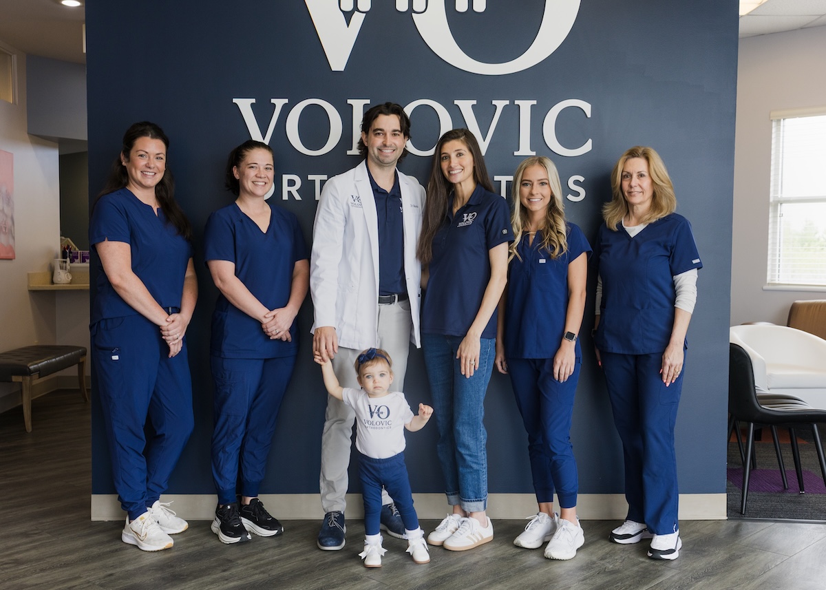 A team of six dental professionals, five women in navy blue scrubs and one man in a white lab coat, pose in front of a navy blue wall with the "VOLOVIC ORTHODONTICS" logo. A young child in a matching Volovic t-shirt and jeans stands in the center foreground, holding the hand of the man in the lab coat. They are all smiling inside a modern, bright office.