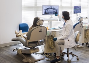 An orthodontist in a white lab coat and blue gloves points to a digital dental X-ray on a computer monitor. He is explaining the scan to a patient who is seated in a grey orthodontic chair. They are in a bright, modern clinic with large windows and multiple treatment stations.