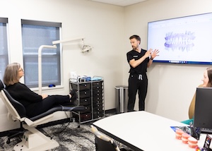 An orthodontist in black scrubs stands in a consultation room, gesturing toward a large television screen displaying a digital scan. A patient sits in a white treatment chair to the left, listening. A table with orthodontic models is in the foreground, and a medical cart is visible against the wall.