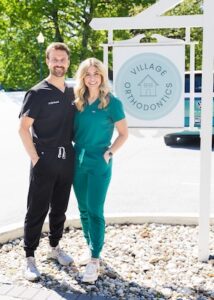 A smiling man and woman stand together in front of an outdoor sign for Village Orthodontics. The man wears black scrubs and the woman wears teal scrubs. They are standing on a gravel area next to a white signpost on a sunny day with green trees in the background.