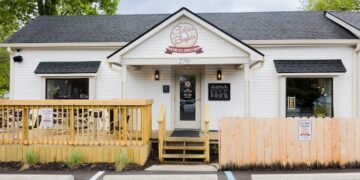 An exterior shot of Gables Bagels, a white-sided building with a black shingled roof. A circular logo above the entrance reads "Gables Bagels" with a banner saying "New York Taste - Zionsville Baked." There is a small wooden deck to the left with outdoor seating, a short wooden fence to the right, and designated customer parking spaces in front.