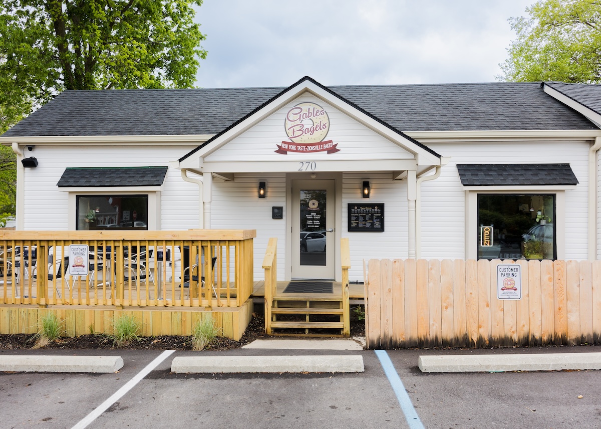 An exterior shot of Gables Bagels, a white-sided building with a black shingled roof. A circular logo above the entrance reads "Gables Bagels" with a banner saying "New York Taste - Zionsville Baked." There is a small wooden deck to the left with outdoor seating, a short wooden fence to the right, and designated customer parking spaces in front.
