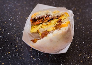 A close-up of a breakfast bagel sandwich wrapped in white deli paper and served in a white paper tray. The sandwich is sliced in half, revealing layers of crispy bacon, a folded egg patty, and melted cheese on a plain bagel. The tray sits on a dark, speckled countertop.