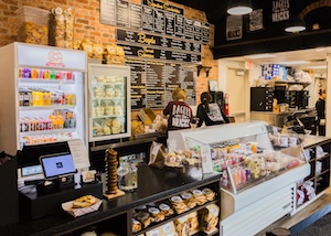 he interior of Gables Bagels showing the ordering counter and a large black menu board on a brick wall. A staff member in a "Bagels & Bricks" t-shirt stands behind the counter. To the left, a refrigerated case is stocked with colorful drinks, and to the right, a glass display case holds various cream cheeses and spreads.