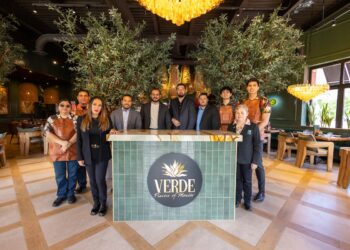 A wide-angle group portrait shows the professional staff of "Verde: Flavors of Mexico" posing in their elegantly designed restaurant. Ten employees, a mix of men and women, are arranged behind and around a central green-tiled host stand that features the restaurant's logo—a stylized agave plant. Some staff members wear leather aprons, while others are in blazers or floral shirts. The interior is upscale and warm, featuring two large indoor olive trees, golden tiered chandeliers, and modern light wood furniture.