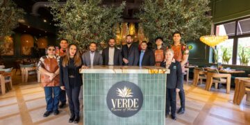A wide-angle group portrait shows the professional staff of "Verde: Flavors of Mexico" posing in their elegantly designed restaurant. Ten employees, a mix of men and women, are arranged behind and around a central green-tiled host stand that features the restaurant's logo—a stylized agave plant. Some staff members wear leather aprons, while others are in blazers or floral shirts. The interior is upscale and warm, featuring two large indoor olive trees, golden tiered chandeliers, and modern light wood furniture.