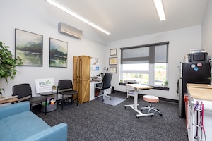 A wide view of a clean, brightly lit treatment room at Inspire Allergy and Asthma. The room features a blue sofa, black chairs, and a desk area. A "inspire" logo sign is on a side table. To the right, there is a mobile medical workstation with a pink stool, a black refrigerator, and cabinets. Landscape artwork and framed certificates decorate the white walls.