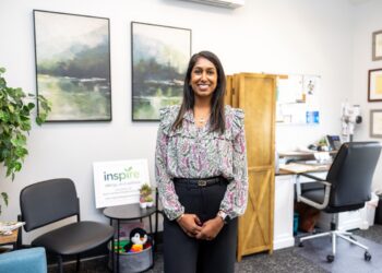 A medium shot of a smiling woman with dark hair standing in a professional office. She is wearing a floral-patterned blouse with ruffle details and black trousers. In the background, there are two landscape paintings, a wooden screen, and a desk. To her left, a small sign reads "inspire allergy and asthma," next to a small shelf with toys and plants.
