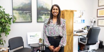 A medium shot of a smiling woman with dark hair standing in a professional office. She is wearing a floral-patterned blouse with ruffle details and black trousers. In the background, there are two landscape paintings, a wooden screen, and a desk. To her left, a small sign reads "inspire allergy and asthma," next to a small shelf with toys and plants.