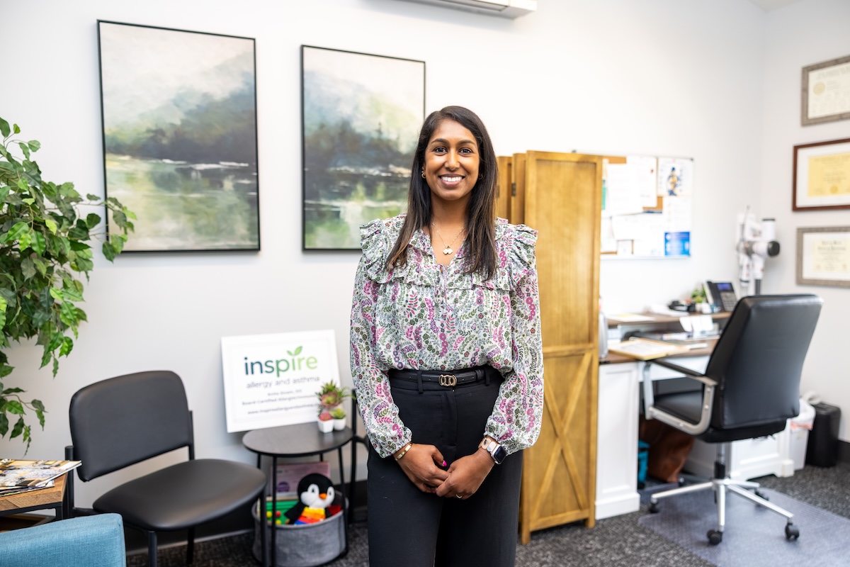 A medium shot of a smiling woman with dark hair standing in a professional office. She is wearing a floral-patterned blouse with ruffle details and black trousers. In the background, there are two landscape paintings, a wooden screen, and a desk. To her left, a small sign reads "inspire allergy and asthma," next to a small shelf with toys and plants.