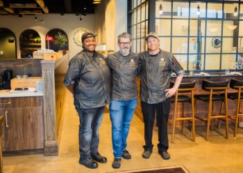 A medium shot of three smiling chefs standing side-by-side in a modern restaurant. The chef on the left is a Black woman in a grey coat and black cap. The chef in the center is a man with a grey beard and glasses in a grey coat and blue jeans. The chef on the right is a man in a grey coat, black pants, and a tan cap. They are standing in front of a bar area with wooden stools and a large glass window.