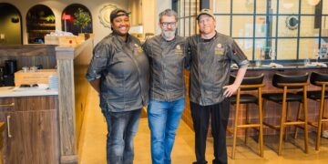 A medium shot of three smiling chefs standing side-by-side in a modern restaurant. The chef on the left is a Black woman in a grey coat and black cap. The chef in the center is a man with a grey beard and glasses in a grey coat and blue jeans. The chef on the right is a man in a grey coat, black pants, and a tan cap. They are standing in front of a bar area with wooden stools and a large glass window.