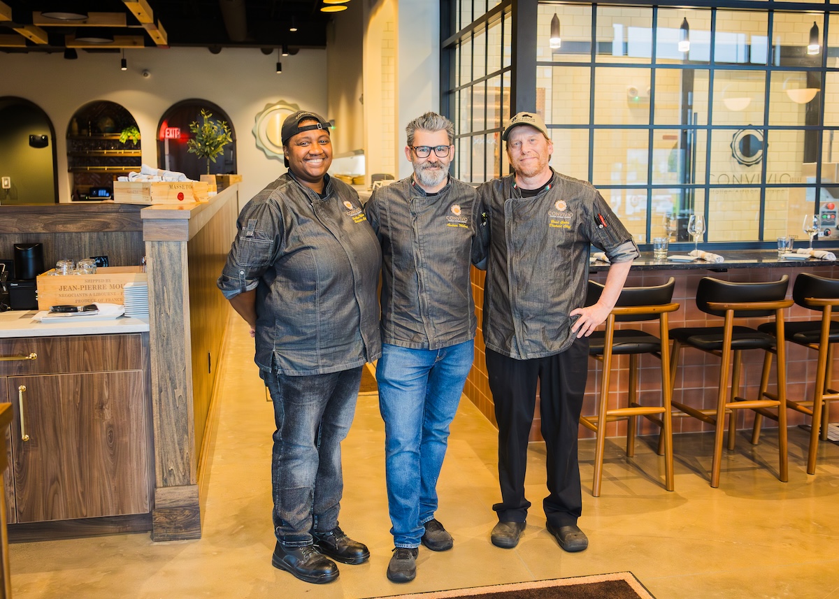 A medium shot of three smiling chefs standing side-by-side in a modern restaurant. The chef on the left is a Black woman in a grey coat and black cap. The chef in the center is a man with a grey beard and glasses in a grey coat and blue jeans. The chef on the right is a man in a grey coat, black pants, and a tan cap. They are standing in front of a bar area with wooden stools and a large glass window.