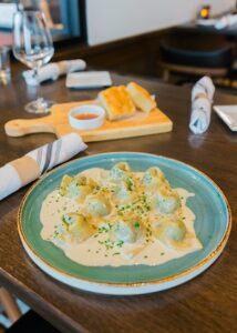 A close-up of a plate of stuffed pasta, likely ravioli or tortellini, covered in a creamy white sauce and garnished with fresh herbs. The dish is served on a teal plate. In the background, a small wooden board holds pieces of focaccia bread and a dipping sauce. A wine glass and rolled napkins sit on the dark wooden table.