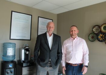 Two men stand smiling in a professional office waiting area. The man on the left is bald, wearing a dark grey blazer over a white shirt. The man on the right has short dark hair and a beard, wearing a light pink button-down shirt and blue trousers. Behind them, framed informational posters about hearing loss hang on a beige wall, next to a water cooler and coffee station. Colorful decorative plates are mounted on the wall to the right.
