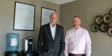 Two men stand smiling in a professional office waiting area. The man on the left is bald, wearing a dark grey blazer over a white shirt. The man on the right has short dark hair and a beard, wearing a light pink button-down shirt and blue trousers. Behind them, framed informational posters about hearing loss hang on a beige wall, next to a water cooler and coffee station. Colorful decorative plates are mounted on the wall to the right.