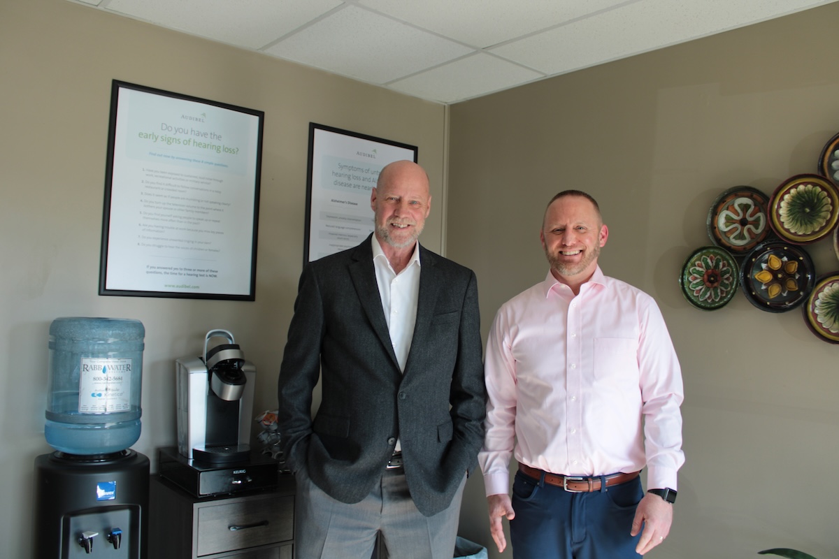 Two men stand smiling in a professional office waiting area. The man on the left is bald, wearing a dark grey blazer over a white shirt. The man on the right has short dark hair and a beard, wearing a light pink button-down shirt and blue trousers. Behind them, framed informational posters about hearing loss hang on a beige wall, next to a water cooler and coffee station. Colorful decorative plates are mounted on the wall to the right.