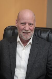A professional headshot of a bald man with a short grey beard, smiling warmly. He is wearing a dark grey blazer over a white button-down shirt. He is seated in a black leather office chair against a solid, warm orange-colored wall.
