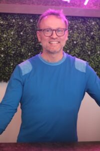 A medium shot of a smiling man with glasses and short hair. He is wearing a blue long-sleeved athletic shirt with grey shoulder patches. He is standing in front of a green foliage wall with a pink neon light visible at the top of the frame.