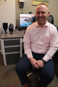 A man with short dark hair and a beard sits in a black mesh office chair, smiling broadly. He is wearing a light pink long-sleeved button-down shirt and dark blue trousers. He is seated next to a grey and white desk with a computer monitor, a small black speaker, and specialized medical tools. The office has neutral-toned walls and a carpeted floor.
