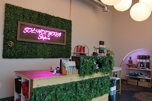 An interior view of the reception area at Sol Hot Yoga. A wooden front desk is decorated with faux greenery. Behind the desk, a pink neon sign reading "SOL HOT YOGA" is mounted on a large green foliage wall. To the right, globe pendant lights hang from the ceiling, and a retail shelf displays yoga-related products.