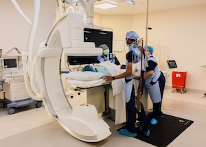 A bright, high-tech interventional radiology suite. A medical team in blue scrubs, surgical caps, and protective lead aprons is gathered around a patient on a treatment table. A large, white C-arm imaging machine curves over the table. Monitors displaying medical data are visible in the background of the clean, sterile room.