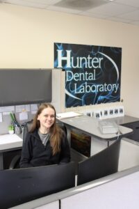 A young woman with long brown hair smiles from behind a reception desk at Hunter Dental Laboratory. She is wearing a black quarter-zip pullover. A large blue and black company sign hangs on the wall behind her, and two computer monitors are visible in the foreground.