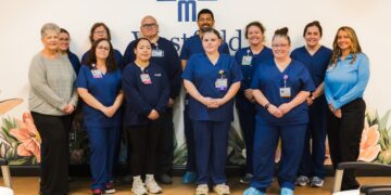 A large group of healthcare professionals posing together in a medical facility. Ten staff members are wearing navy blue scrubs, one woman on the far left is in a grey sweater, and one woman on the far right is in a light blue polo. They are standing in front of a white wall with a blue geometric logo and a floral mural.