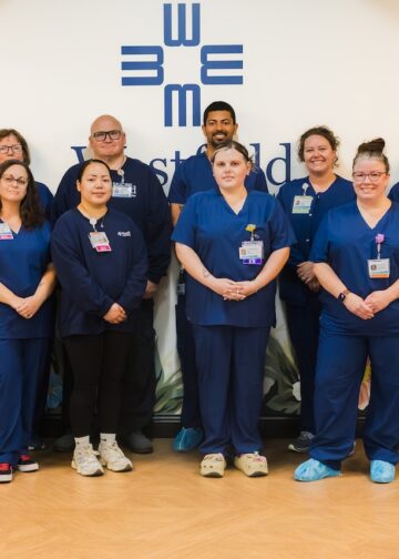 A large group of healthcare professionals posing together in a medical facility. Ten staff members are wearing navy blue scrubs, one woman on the far left is in a grey sweater, and one woman on the far right is in a light blue polo. They are standing in front of a white wall with a blue geometric logo and a floral mural.