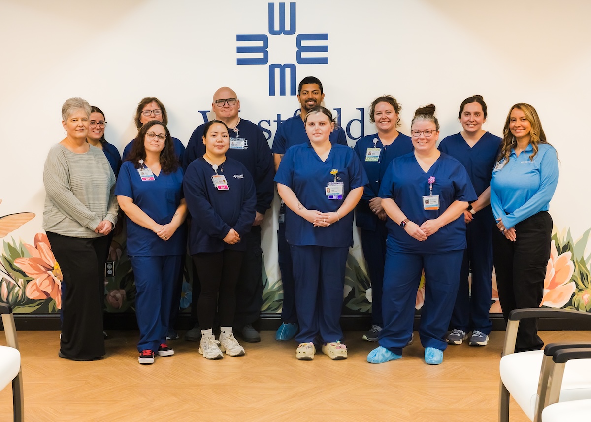 A large group of healthcare professionals posing together in a medical facility. Ten staff members are wearing navy blue scrubs, one woman on the far left is in a grey sweater, and one woman on the far right is in a light blue polo. They are standing in front of a white wall with a blue geometric logo and a floral mural.
