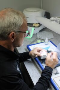 A dental technician in a black long-sleeve shirt and glasses works meticulously on a white dental model at a lab bench. He uses a small tool to refine the mold. In the background, specialized equipment, including a dental vacuum mixer, is visible under bright task lighting