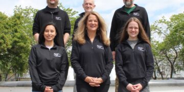 Six members of the Hunter Dental Laboratory team stand together for a group photo outdoors. They are arranged on concrete steps with a fountain and green trees in the background. The team is wearing matching black quarter-zip pullovers with the company logo on the chest.
