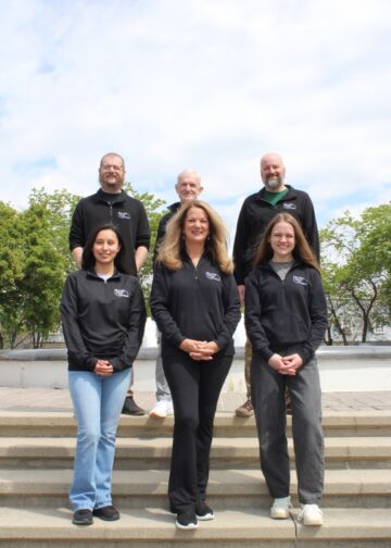Six members of the Hunter Dental Laboratory team stand together for a group photo outdoors. They are arranged on concrete steps with a fountain and green trees in the background. The team is wearing matching black quarter-zip pullovers with the company logo on the chest.