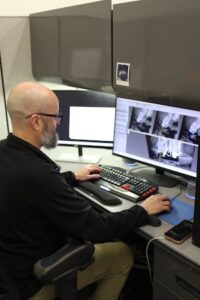 A technician with a grey beard and glasses sits at a desk, reviewing dental X-rays on a computer monitor. He is using a keyboard and mouse to analyze the digital images, which show detailed views of teeth and jaw structures. The office setting includes a secondary monitor and grey privacy partitions. 