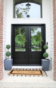 A close-up shot of a modern home's front entryway. The entrance features elegant black double doors with large glass panes, each adorned with a lush green wreath. Two tall, grey planters with double-ball topiaries flank the doors, and a stylish "WELCOME" doormat sits on the concrete porch over a black-and-white striped rug.