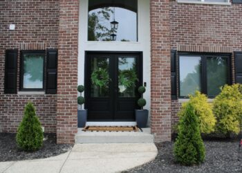 A front entrance of a two-story red brick house features black double doors with green wreaths. Two matching black windows with shutters flank the door. A concrete walkway leads to the entrance, which is decorated with two potted topiary trees and small evergreen shrubs in the mulch.