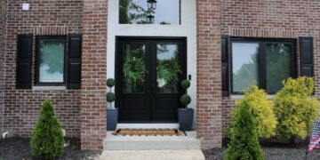 A front entrance of a two-story red brick house features black double doors with green wreaths. Two matching black windows with shutters flank the door. A concrete walkway leads to the entrance, which is decorated with two potted topiary trees and small evergreen shrubs in the mulch.