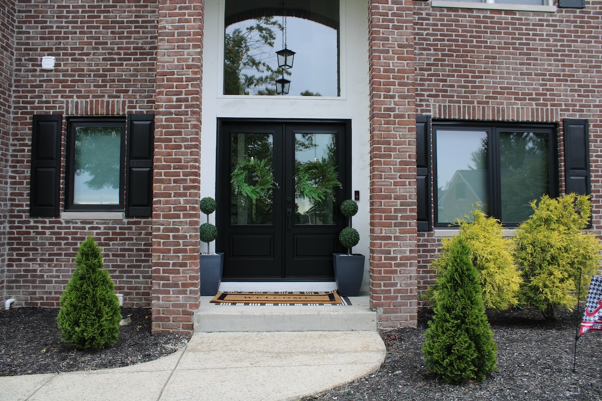A front entrance of a two-story red brick house features black double doors with green wreaths. Two matching black windows with shutters flank the door. A concrete walkway leads to the entrance, which is decorated with two potted topiary trees and small evergreen shrubs in the mulch.