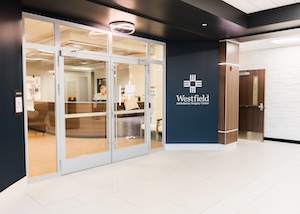 A clean and modern medical office entrance for Riverview Health in Westfield. The entrance features large glass double doors with white frames. To the right of the doors is a navy blue accent wall with the white Riverview Health logo. Through the glass, a brightly lit reception desk and waiting area are visible.