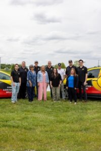 An eye-level group portrait shows 11 employees of INC Concrete & Excavation Solutions standing on a green lawn. The team, a mix of men and women, is dressed in a blend of professional and casual attire, including black polo shirts with the company logo, denim jackets, and bright blouses. They are framed by two red and yellow company vehicles on either side. The background features a wide, grassy field under a bright, overcast sky.