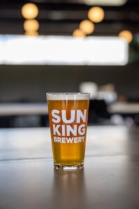 A vertical shot of a pint glass filled with a clear, golden-amber beer, topped with a thin white head. The glass features the "SUN KING BREWERY" logo in bold white letters. It sits on a light-colored wooden table with a blurred brewery interior and warm, glowing globe lights in the background.