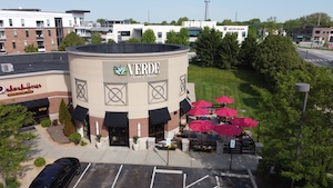 An aerial view shows the exterior of "Verde Flavors of Mexico," a restaurant located in a modern, circular stone building. To the right, a patio area is shaded by several bright pink umbrellas, situated next to a lush green lawn. The building features black awnings over large windows and a sign with the restaurant's logo. In the background, other commercial and residential buildings are visible under a clear, sunny sky.
